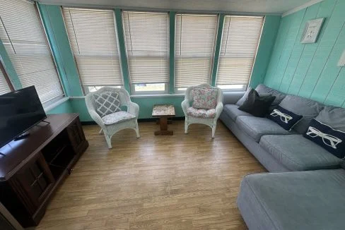 A room with teal walls features a couch, two wicker chairs, a wooden TV stand with a flatscreen TV, and several windows with closed blinds. The floor is wooden.
