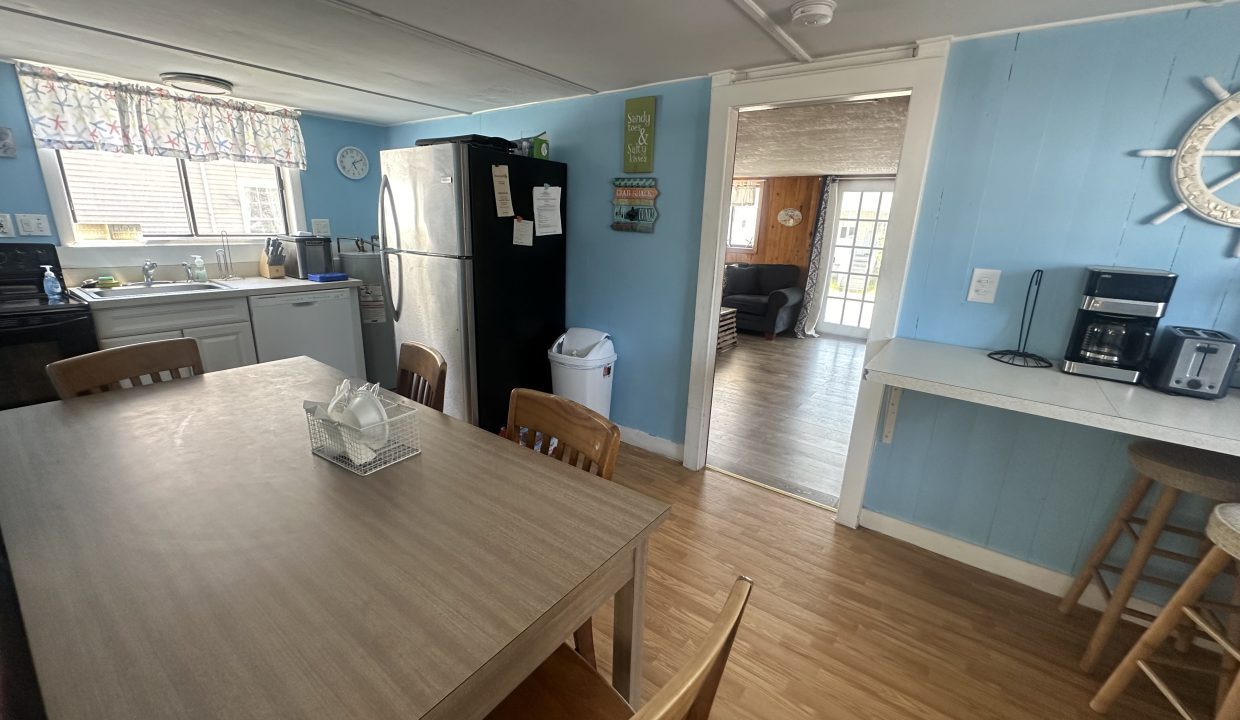 A kitchen with light blue walls featuring a dining table, chairs, stainless steel refrigerator, sink, stove, and a coffee maker.  There's a doorway leading to another room with a couch and sliding glass doors.