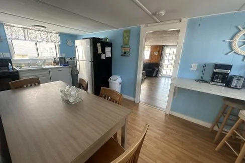 A kitchen with light blue walls featuring a dining table, chairs, stainless steel refrigerator, sink, stove, and a coffee maker.  There's a doorway leading to another room with a couch and sliding glass doors.