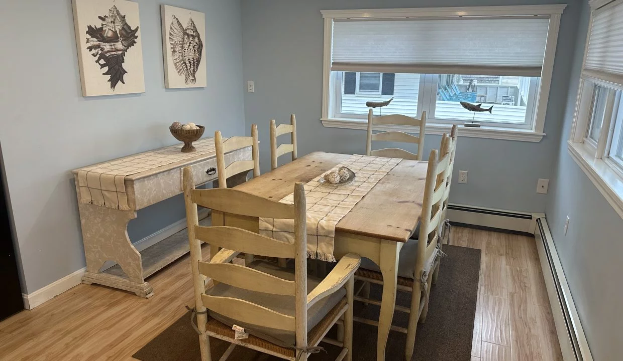 A light blue dining room with a wooden table and six chairs, a side table against the wall, and two framed art pieces of seashells. The room has a wooden floor and sunlight coming through the windows.