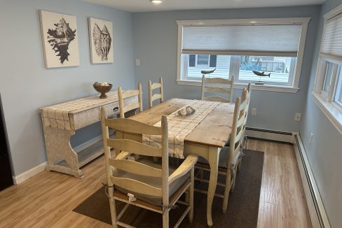 A light blue dining room with a wooden table and six chairs, a side table against the wall, and two framed art pieces of seashells. The room has a wooden floor and sunlight coming through the windows.
