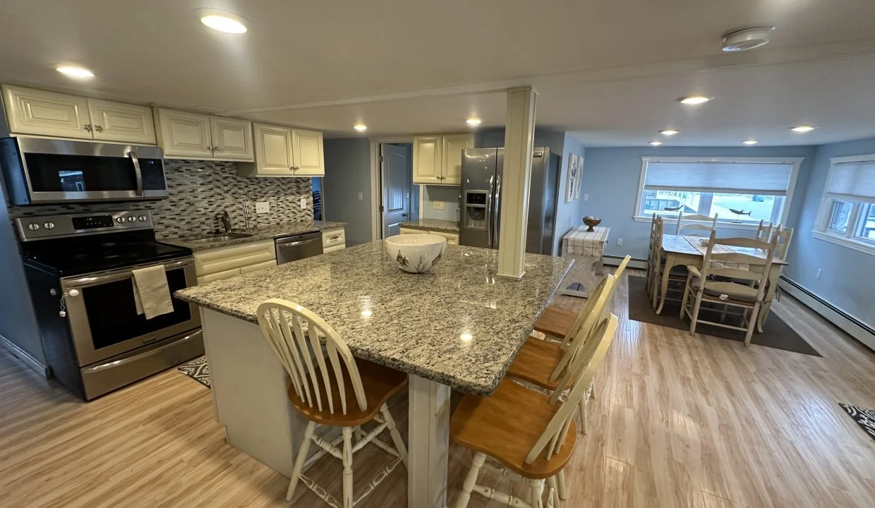 A modern kitchen with granite countertops, white cabinets, stainless steel appliances, and wooden stools at the island. A dining table with white chairs is in the background. The floor has light wood planks.