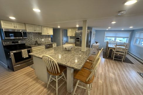 A modern kitchen with granite countertops, white cabinets, stainless steel appliances, and wooden stools at the island. A dining table with white chairs is in the background. The floor has light wood planks.