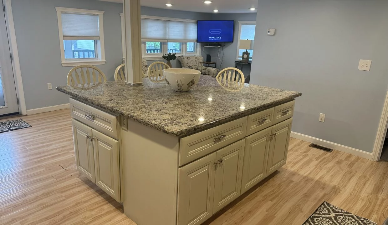 A kitchen with light wooden flooring, a granite-topped island, white cabinets, and cushioned chairs. In the background, there is a TV on a stand and windows with shades.