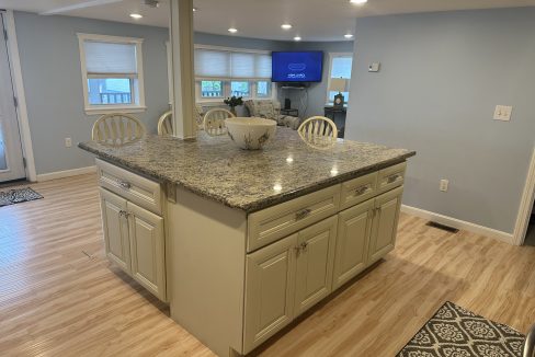 A kitchen with light wooden flooring, a granite-topped island, white cabinets, and cushioned chairs. In the background, there is a TV on a stand and windows with shades.