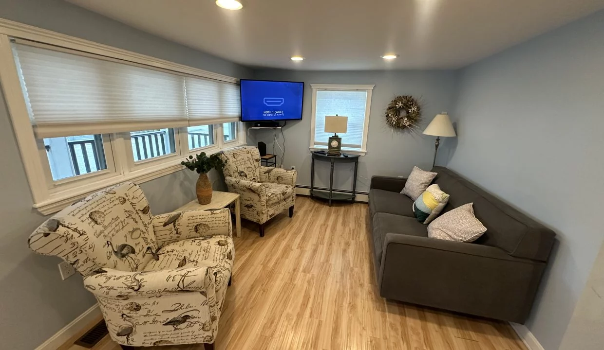 A small living room with light blue walls, featuring two armchairs with patterned fabric, a gray couch, a wall-mounted television, a small table with a lamp, and a wreath hanging on the wall.