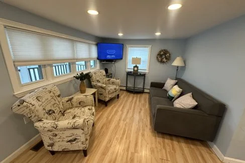 A small living room with light blue walls, featuring two armchairs with patterned fabric, a gray couch, a wall-mounted television, a small table with a lamp, and a wreath hanging on the wall.