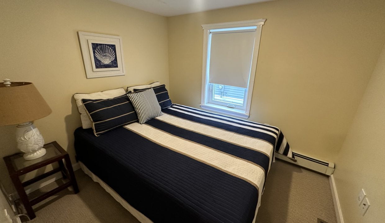 A small bedroom with a window, a bed fitted with navy and white striped bedding, a bedside table with a lamp, a framed picture on the wall, and beige walls and carpet.