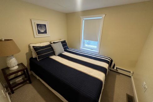 A small bedroom with a window, a bed fitted with navy and white striped bedding, a bedside table with a lamp, a framed picture on the wall, and beige walls and carpet.