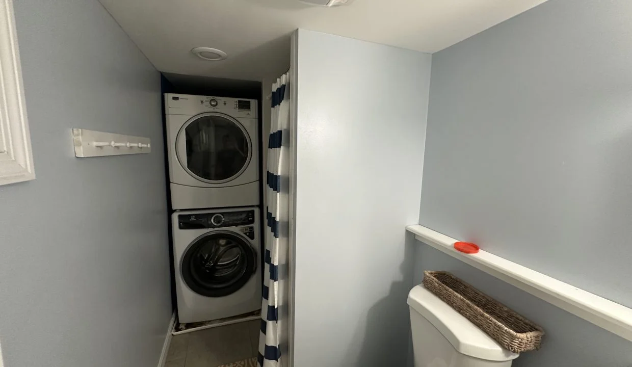 A small laundry room with a stacked washer and dryer, a striped shower curtain, and a toilet. There is a wall-mounted hook and a wicker basket on the shelf above the toilet.