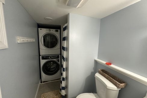 A small laundry room with a stacked washer and dryer, a striped shower curtain, and a toilet. There is a wall-mounted hook and a wicker basket on the shelf above the toilet.