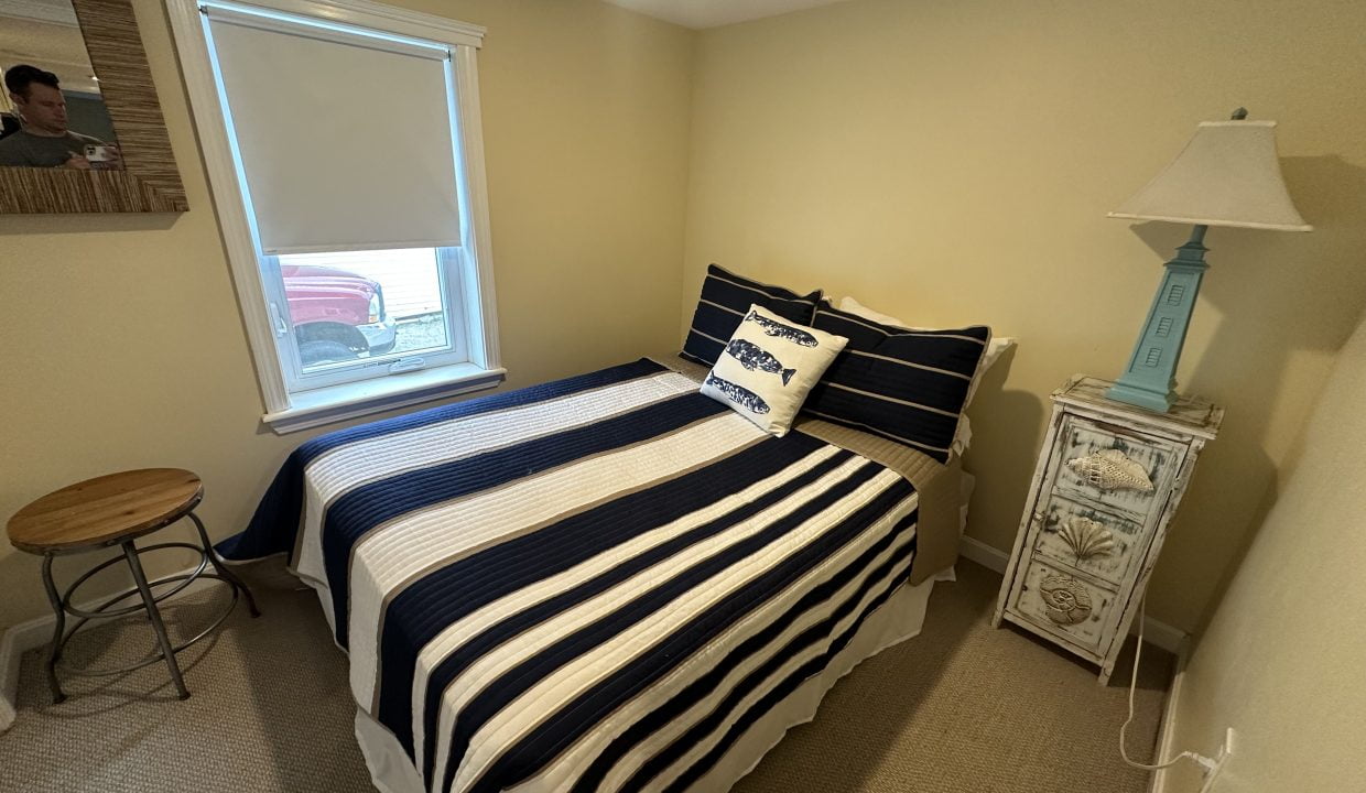 Small bedroom with a neatly made bed featuring striped bedding. A nightstand with a blue lamp and seashell decorations sits next to the bed. There's a stool by a window with a blind, and a wall picture.