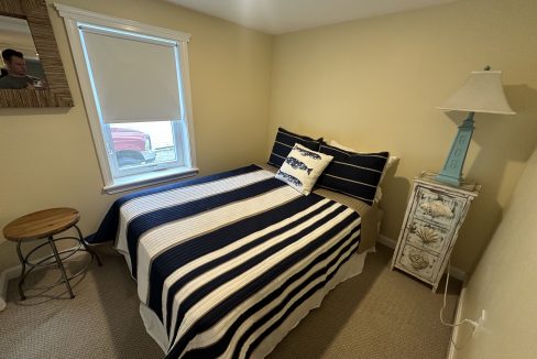 Small bedroom with a neatly made bed featuring striped bedding. A nightstand with a blue lamp and seashell decorations sits next to the bed. There's a stool by a window with a blind, and a wall picture.