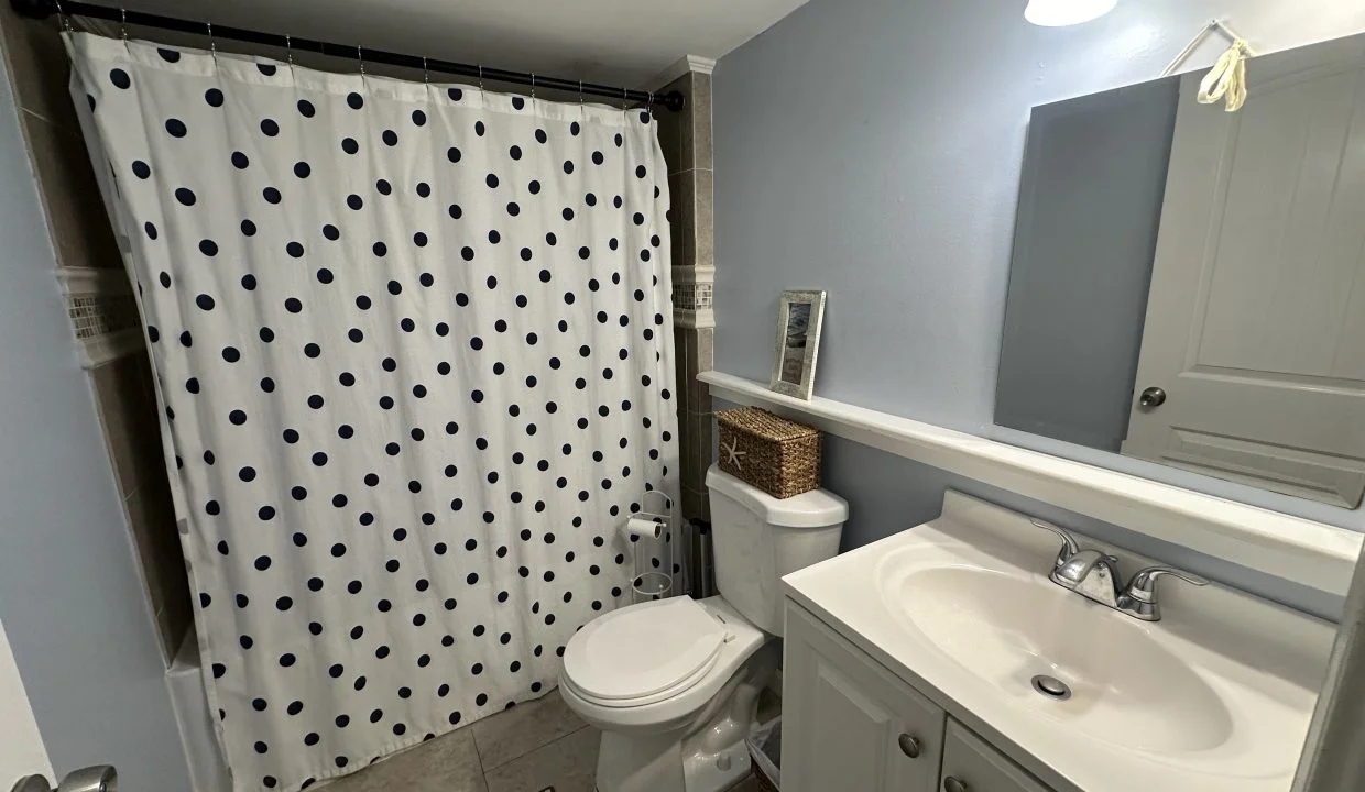 A bathroom with a polka dot shower curtain, a toilet, a white sink with a cabinet, a mirror, a wicker basket, and a small picture frame on the toilet tank. A light fixture is above the sink.