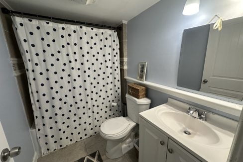 A bathroom with a polka dot shower curtain, a toilet, a white sink with a cabinet, a mirror, a wicker basket, and a small picture frame on the toilet tank. A light fixture is above the sink.