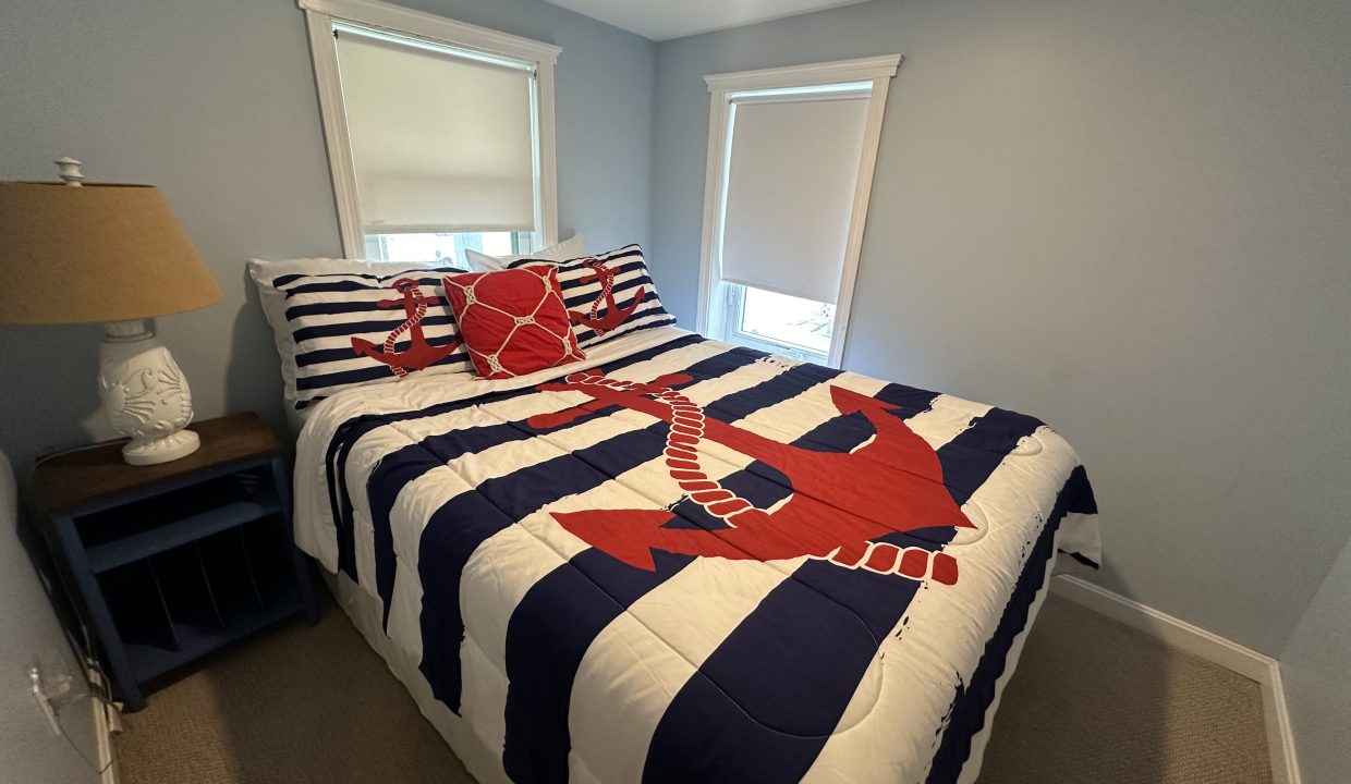 Small bedroom with two windows, a bed with a nautical-themed comforter featuring an anchor and crab, a nightstand with a lamp, and light-colored walls and flooring.