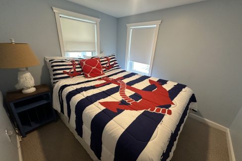 Small bedroom with two windows, a bed with a nautical-themed comforter featuring an anchor and crab, a nightstand with a lamp, and light-colored walls and flooring.