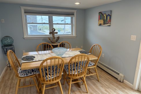 A dining room with a wooden table set for six. The table is decorated with a blue centerpiece and placemats. The room has light blue walls, wooden chairs, and a wall painting.