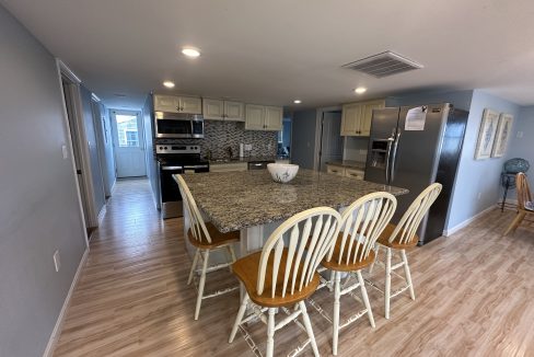 A modern kitchen with a large granite island, four chairs, stainless steel appliances, and wooden floor. A hallway is visible on the left and a dining area on the right.