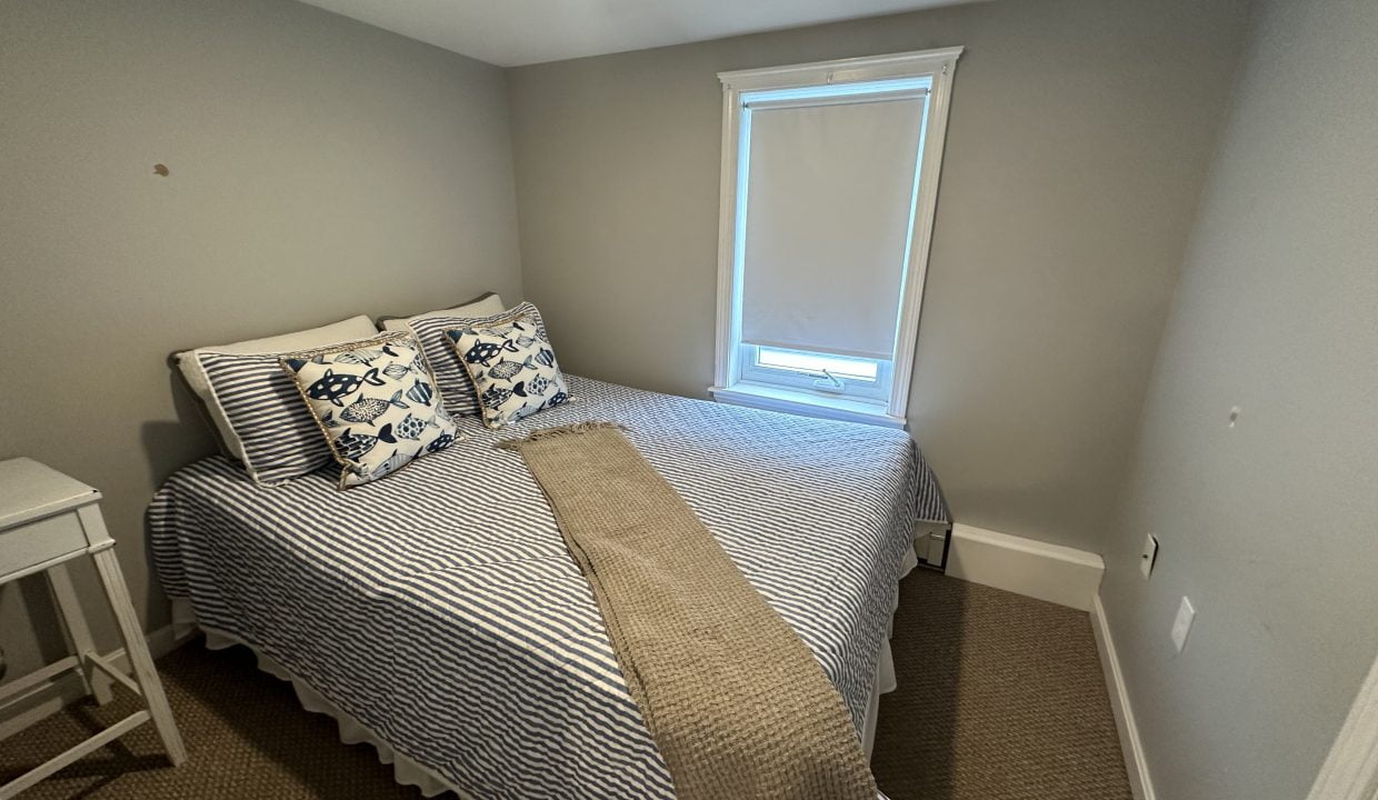 Small bedroom with a single window, a bed with striped bedding, two patterned pillows, and a beige throw. A white nightstand is placed beside the bed. Walls are painted light grey.