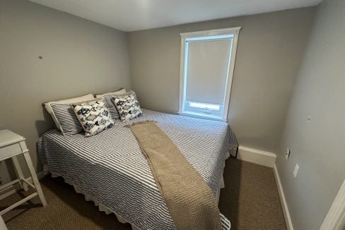 Small bedroom with a single window, a bed with striped bedding, two patterned pillows, and a beige throw. A white nightstand is placed beside the bed. Walls are painted light grey.
