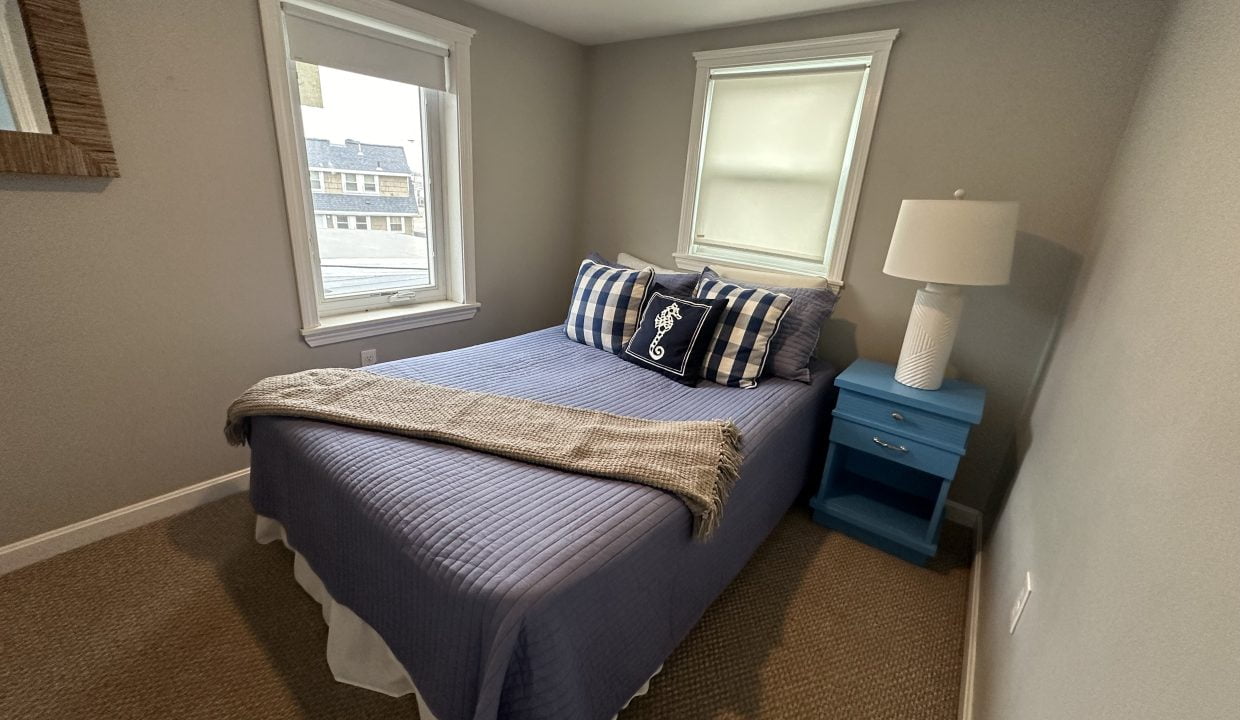 A small bedroom with a double bed covered in a blue quilt and plaid pillows, a blue nightstand with a white lamp, two windows, a wall-mounted mirror, and beige carpeted flooring.