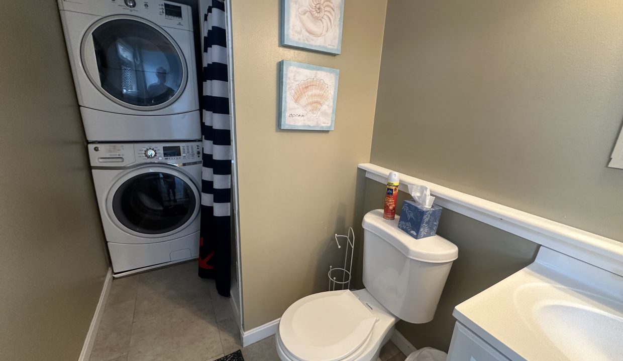 A small bathroom with a toilet, sink, stacked washer and dryer, striped shower curtain, and two seashell-themed wall decorations.