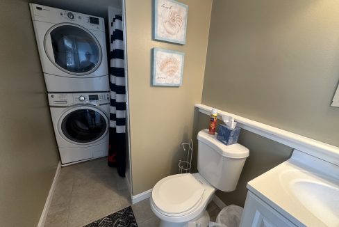 A small bathroom with a toilet, sink, stacked washer and dryer, striped shower curtain, and two seashell-themed wall decorations.