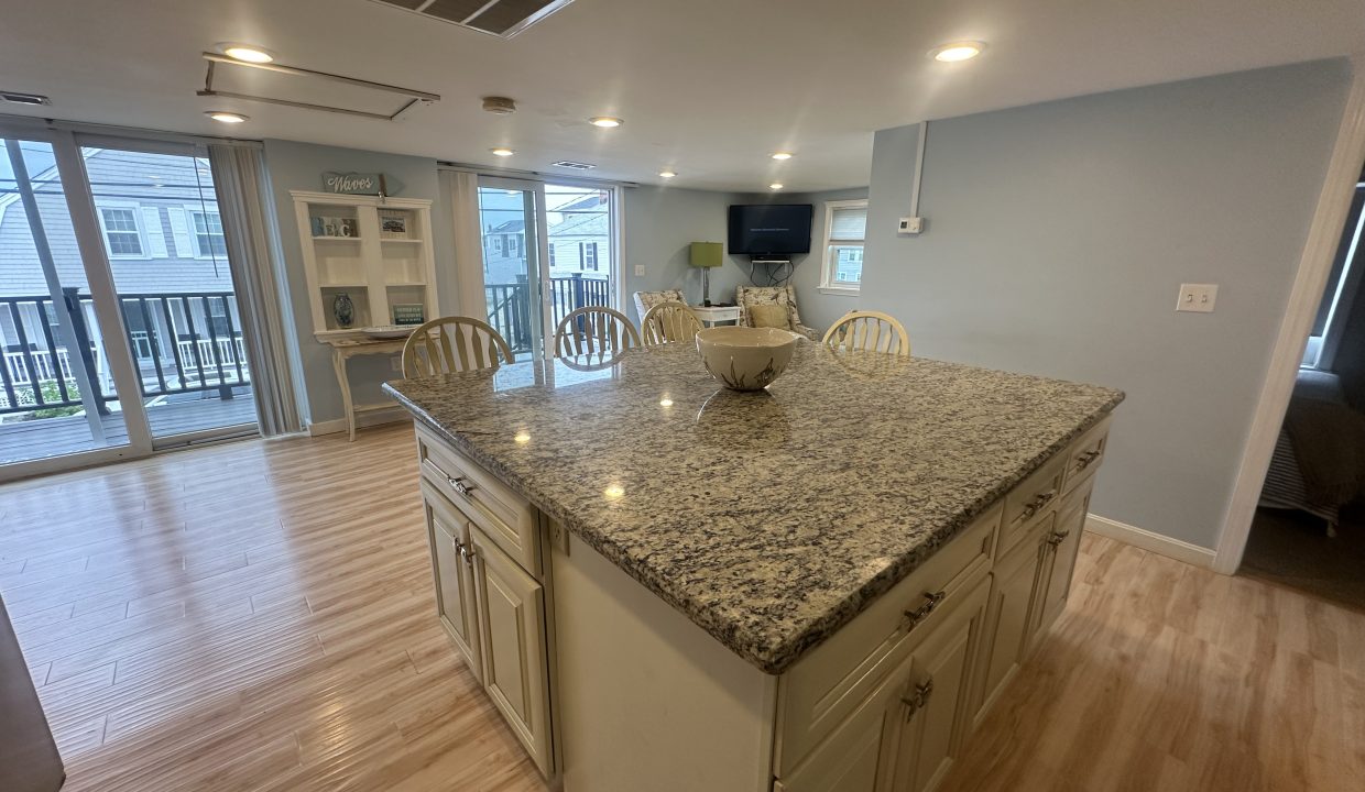 A kitchen with a large granite island, surrounded by stools, and a cabinet in the background. The area is bright with natural light coming through large sliding glass doors that lead to a balcony.