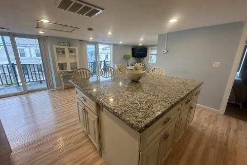 A kitchen with a large granite island, surrounded by stools, and a cabinet in the background. The area is bright with natural light coming through large sliding glass doors that lead to a balcony.