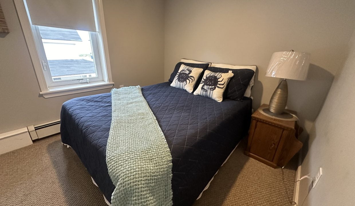 A bedroom with a double bed featuring a dark blue quilt, two pillows with lobster designs, a light blue throw, a wooden bedside table with a lamp, and a window with a blind.