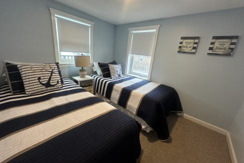 A small bedroom with twin beds, navy and white striped bedding, two windows with white blinds, and ocean-themed wall decor. A lamp and anchor-themed pillows are on one of the beds.
