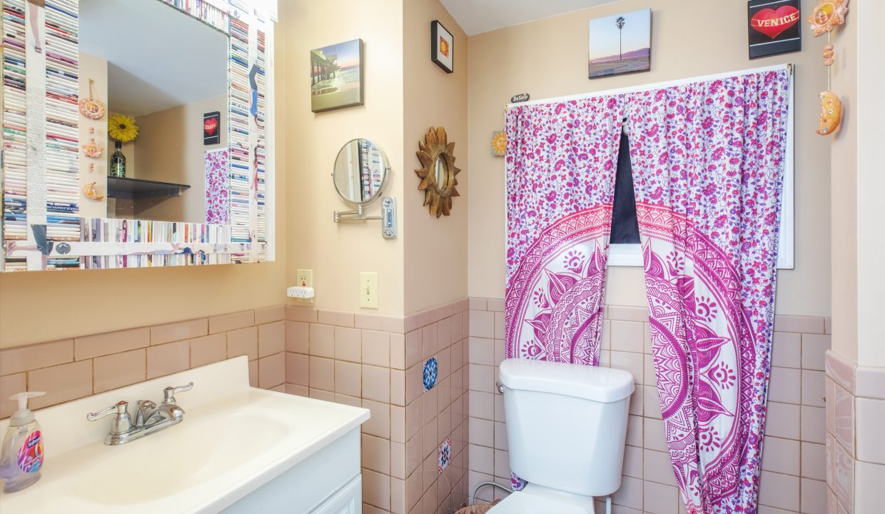 A bathroom with a sink and toilet, decorated with a pink patterned curtain and various framed pictures on light-colored walls. The sink has a mirror above it and a soap dispenser on the counter.