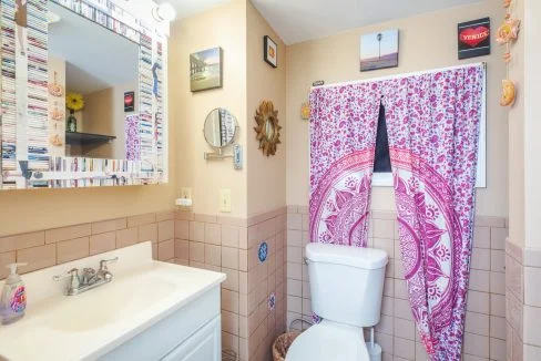 A bathroom with a sink and toilet, decorated with a pink patterned curtain and various framed pictures on light-colored walls. The sink has a mirror above it and a soap dispenser on the counter.