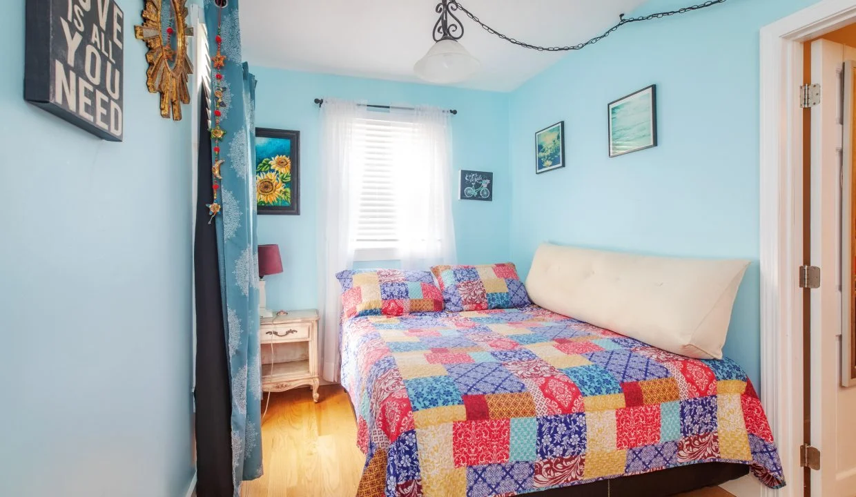 A bright bedroom with light blue walls, a bed with a colorful patchwork quilt, a wooden nightstand with a lamp, and various wall decorations, including a mirror and framed pictures.