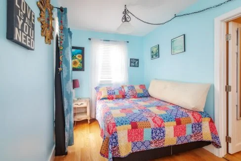 A bright bedroom with light blue walls, a bed with a colorful patchwork quilt, a wooden nightstand with a lamp, and various wall decorations, including a mirror and framed pictures.