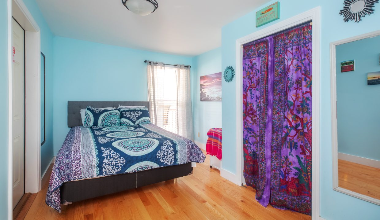 A bedroom with light blue walls, a bed with circular patterned bedding, wooden flooring, a window with blinds, and colorful door curtains.