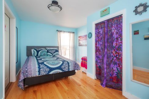 A bedroom with light blue walls, a bed with circular patterned bedding, wooden flooring, a window with blinds, and colorful door curtains.