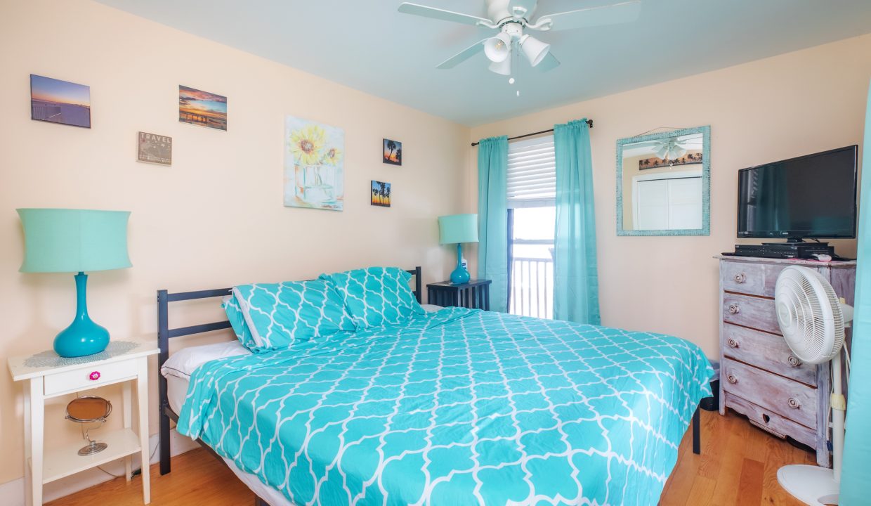 A bedroom with a teal and white patterned bedspread, teal lamps, light pink walls, wall art, a ceiling fan, a window with teal curtains, a white nightstand, a dresser with a TV, and a floor fan.