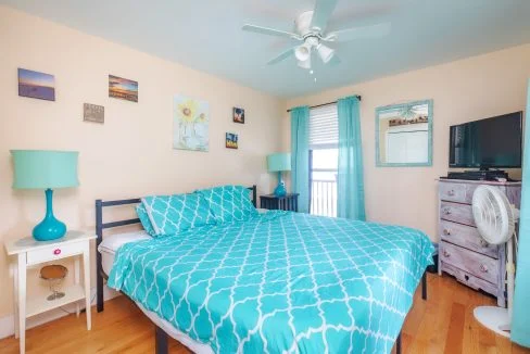 A bedroom with a teal and white patterned bedspread, teal lamps, light pink walls, wall art, a ceiling fan, a window with teal curtains, a white nightstand, a dresser with a TV, and a floor fan.