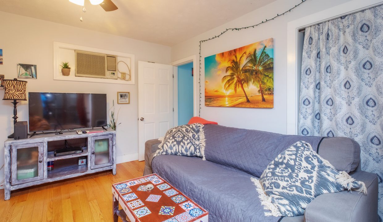 A cozy living room with a gray couch, patterned pillows, a coffee table, a TV, wall art featuring a beach scene, an air conditioner, and a ceiling fan. The room has wooden flooring and light-blue walls.
