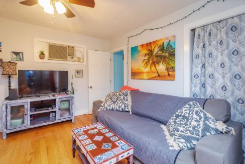 A cozy living room with a gray couch, patterned pillows, a coffee table, a TV, wall art featuring a beach scene, an air conditioner, and a ceiling fan. The room has wooden flooring and light-blue walls.