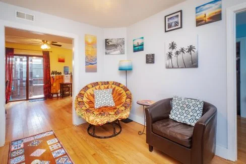 A small, cozy room with a brown leather chair and a circular chair with an orange cushion. The walls are decorated with various framed pictures. The room opens to a larger space with wooden floors.