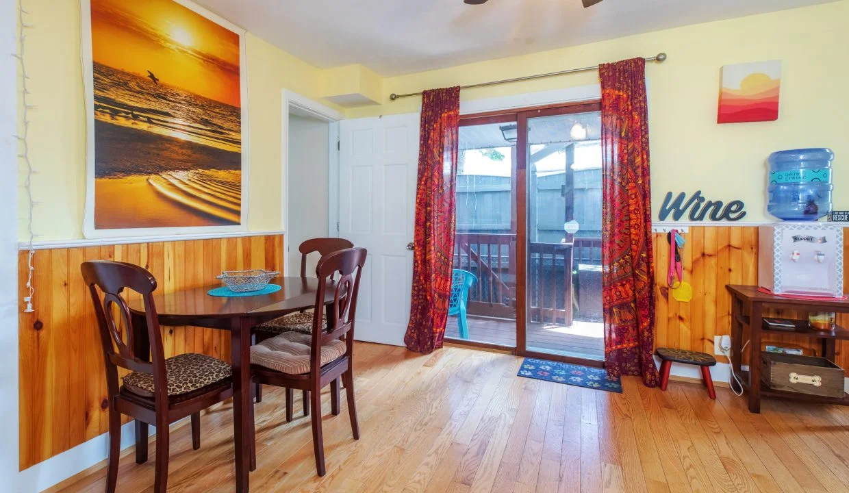 A small dining area with a wooden table and chairs, bright yellow walls, and a sliding glass door. Decor includes a landscape photo, 