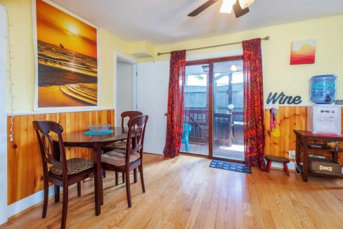 A small dining area with a wooden table and chairs, bright yellow walls, and a sliding glass door. Decor includes a landscape photo, 