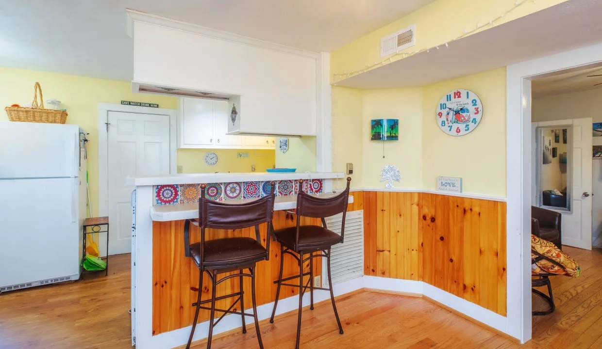 A cozy kitchen with yellow walls and wood paneling, featuring a small bar area with two brown barstools, a clock on the wall, and a white refrigerator in the corner.