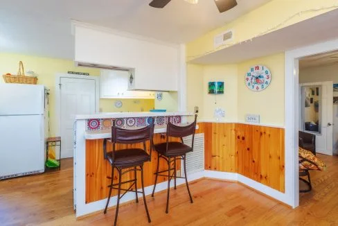 A cozy kitchen with yellow walls and wood paneling, featuring a small bar area with two brown barstools, a clock on the wall, and a white refrigerator in the corner.