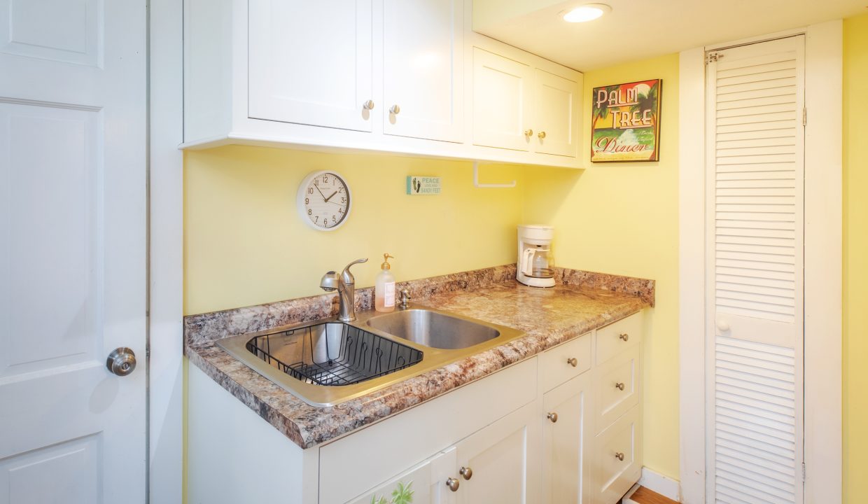 A small kitchen with yellow walls, white cabinets, a marble countertop, a stainless steel sink, a clock, a coffee maker, and a 