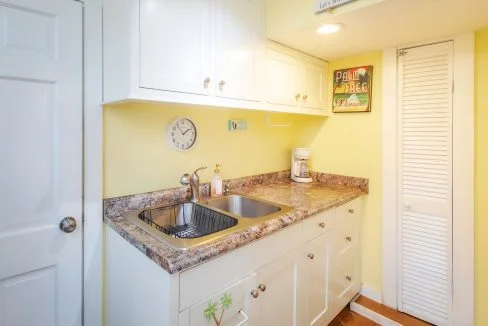 A small kitchen with yellow walls, white cabinets, a marble countertop, a stainless steel sink, a clock, a coffee maker, and a 