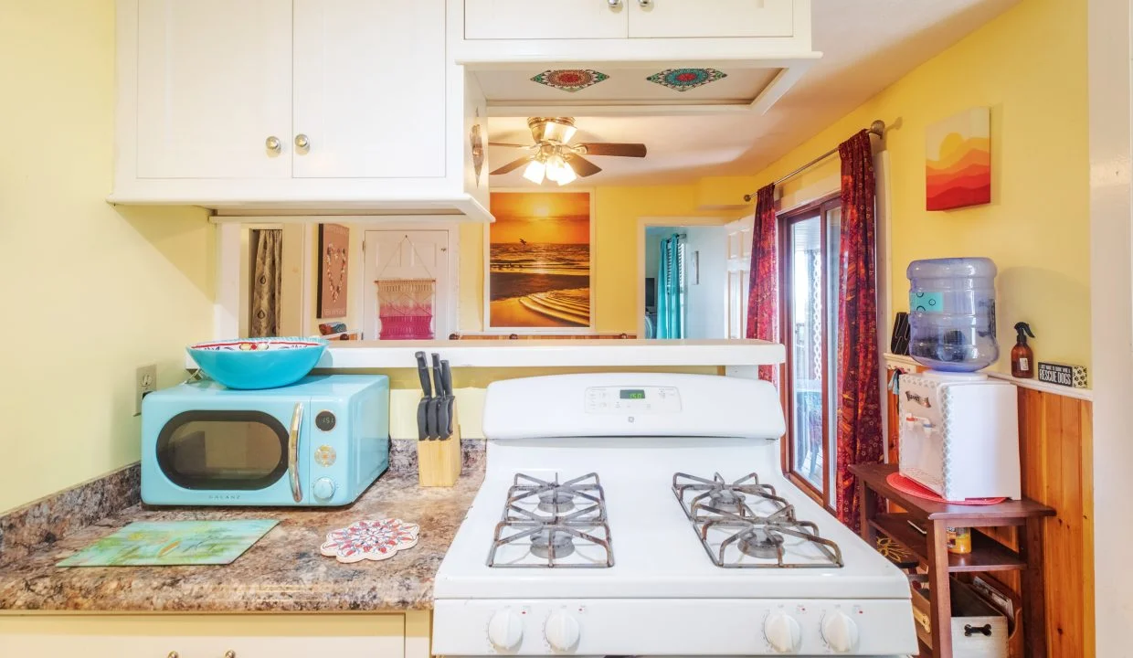 A kitchen with a white gas stove, white cabinets, a microwave oven, a knife block, and a water dispenser. Yellow walls and a ceiling fan in the background.